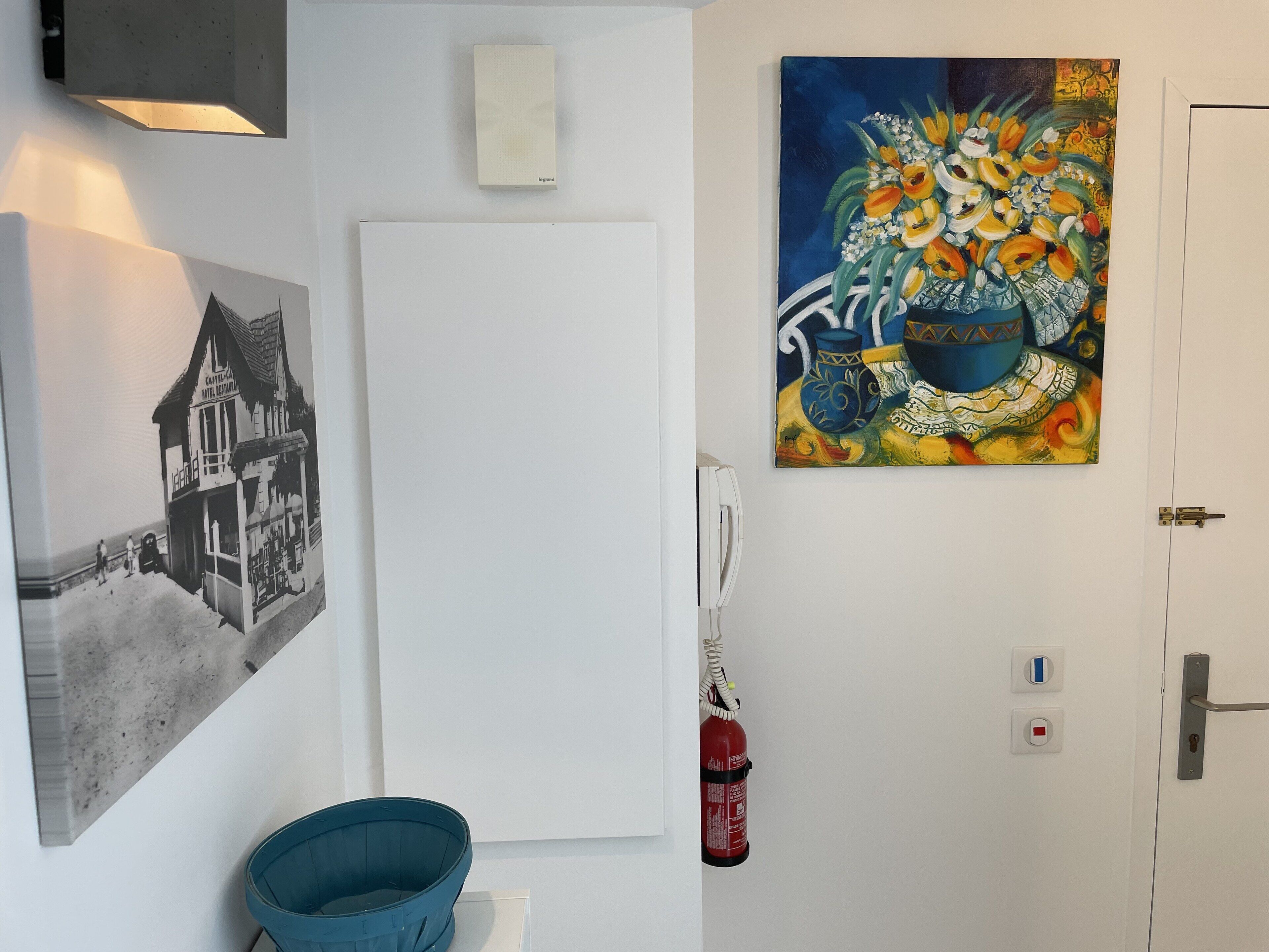A hotel room with a black and white photograph of a building, a colorful painting of flowers, a blue bucket, and a fire extinguisher.