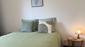 A bedroom with a green bedspread, two pillows, a wall-mounted picture, a bedside lamp, and a small wooden side table. - Apartment in renovated pool villa 2025 (Porto-Vecchio)