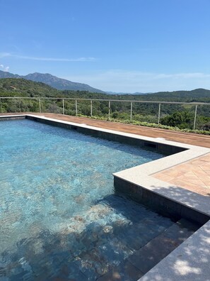 A clear swimming pool with a wooden deck and a metal railing, surrounded by lush greenery and mountains in the background. - Apartment in renovated pool villa 2025 (Porto-Vecchio)