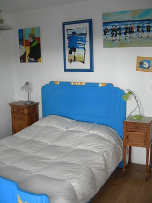 Ein Schlafzimmer mit blauem Kopfteil, einem Bett mit weißer Daunendecke, einem Nachttisch aus Holz mit einer Lampe und Bildern an den Wänden.