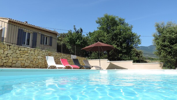 Une piscine entourée de murs de pierre et d’arbres, avec des chaises longues et un parasol.