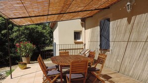 Une terrasse extérieure avec une table et des chaises en bois, un pot de fleurs et une pergola offrant de l’ombre.