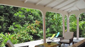 Un patio couvert aménagé avec des meubles en bois, une table recouverte d’une nappe colorée et une végétation tropicale luxuriante, visible à travers les arcades.