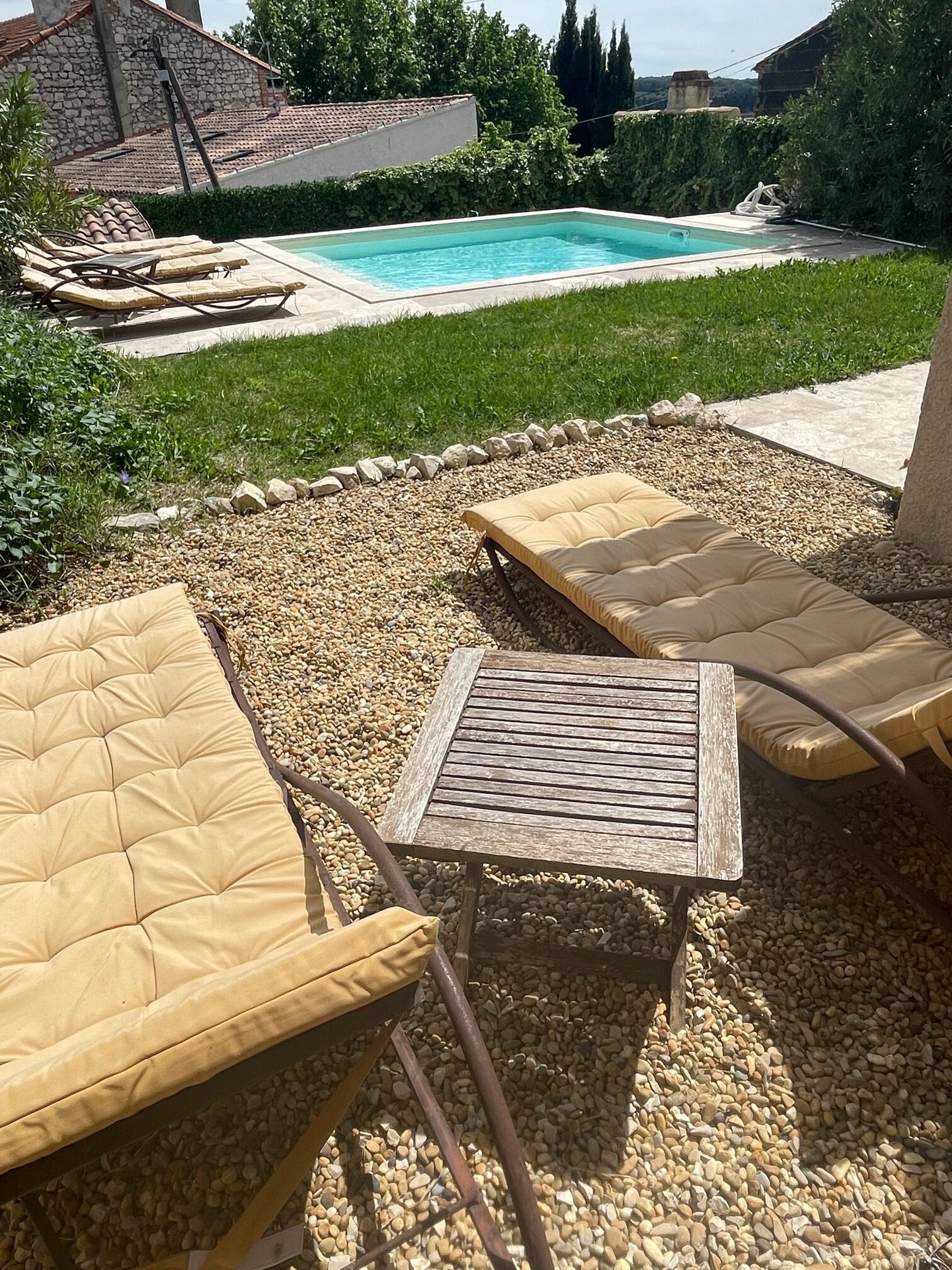 Un espace aménagé au bord de la piscine, avec des chaises longues et une petite table, entouré de galets et de verdure.