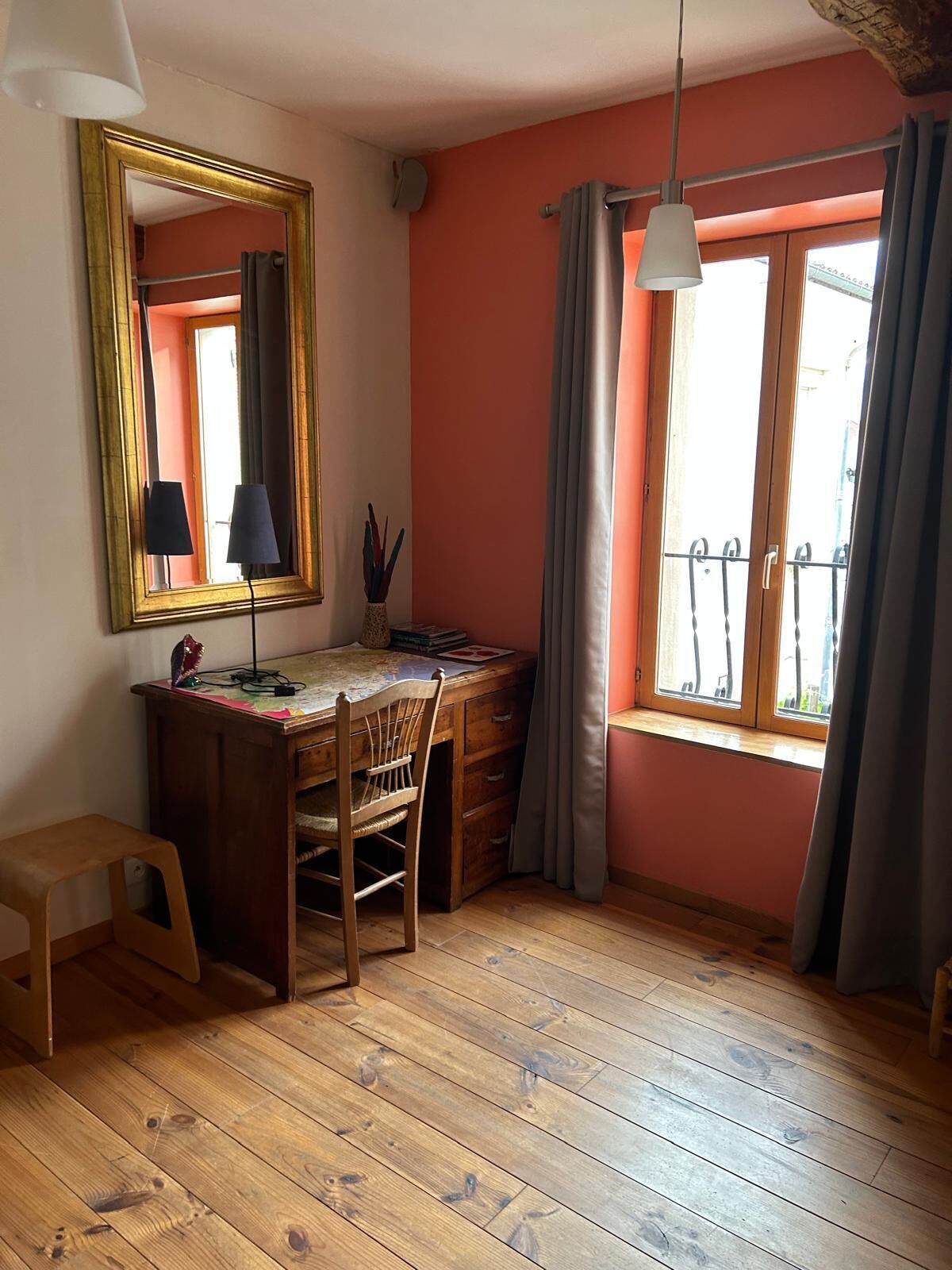 A room with a wooden desk, a chair, a small stool, a mirror, and a window with curtains.