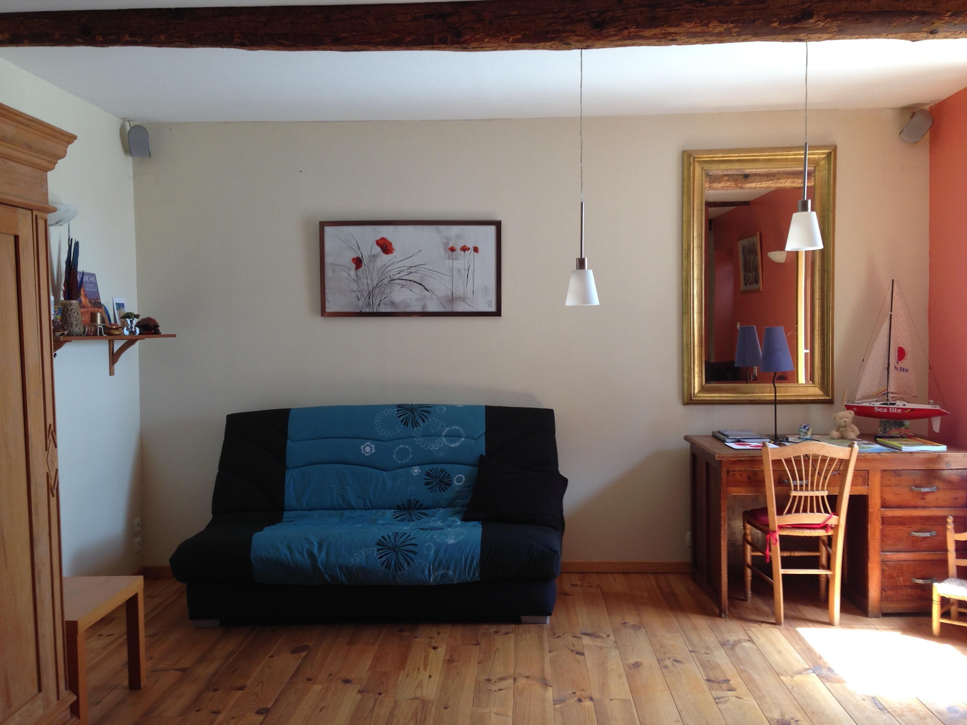 Een knus ingerichte kamer met een houten vloer, een blauwe bank, een houten bureau met stoel, een klein tafeltje en een lijst met een afbeelding aan de muur.