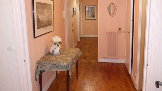 Un pasillo con suelo de madera, una mesa con un florero, cuadros en las paredes y un radiador.