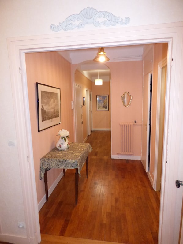 A hallway with wooden flooring, a table with a vase, framed pictures on the walls, and a radiator.