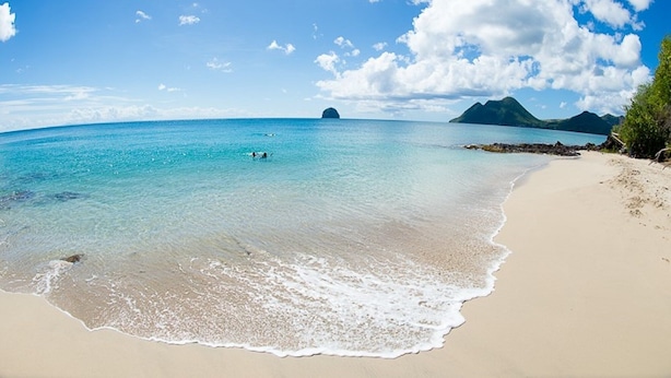 Una playa con agua turquesa cristalina, una orilla arenosa y montañas a lo lejos.