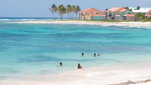 A beach with clear turquoise water, people swimming, and a row of houses with red roofs.