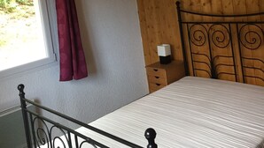 Ett sovrum med ett träpanelat tak, en vit säng med ett metallgavel, ett litet sängbord med en lampa och ett fönster med en röd gardin.