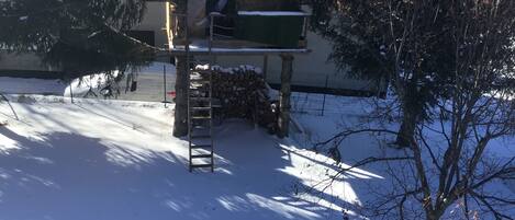 A snow-covered outdoor area with a wooden structure, a ladder, and a pile of firewood.