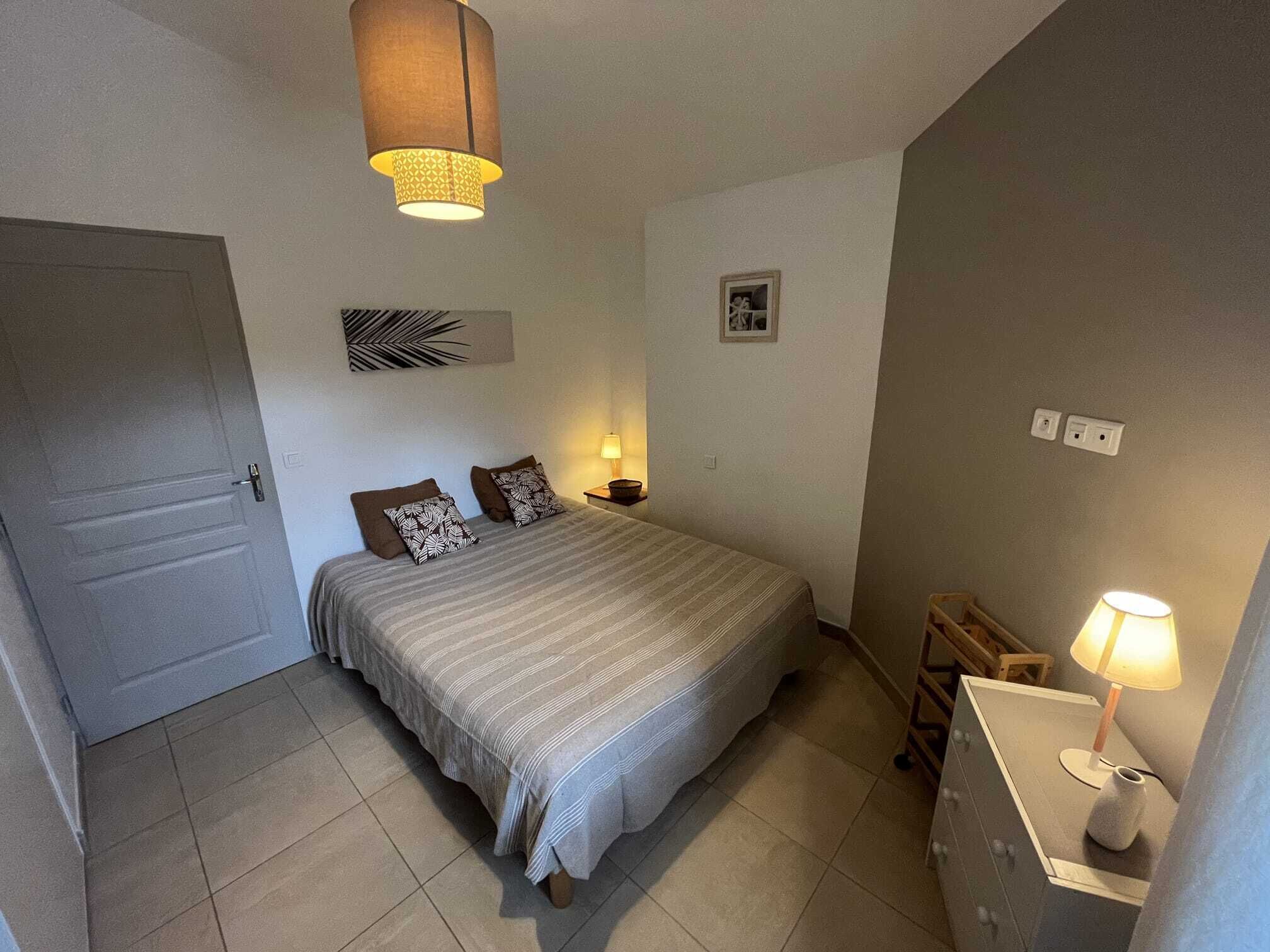 Een slaapkamer met een bed, een nachtkastje, een lamp, een deur en een muur met een klein schilderij.