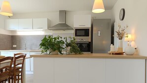 Eine moderne Küche mit weißen Schränken, einer zentralen Kochinsel, einer Edelstahl-Dunstabzugshaube, einer Mikrowelle und einem Backofen. Auf der Insel stehen Blumentöpfe und an der Wand hängt eine Uhr.