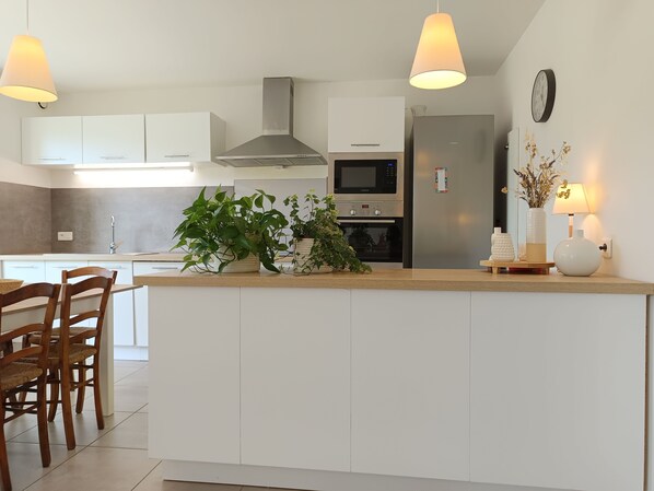 A modern kitchen with white cabinetry, a central island, a stainless steel range hood, a microwave, and an oven. There are potted plants on the island and a clock on the wall. - 9x4 SWIMMING POOL, heated* if needed / Interior fully renovated and modern, natural style  (Ménerbes)