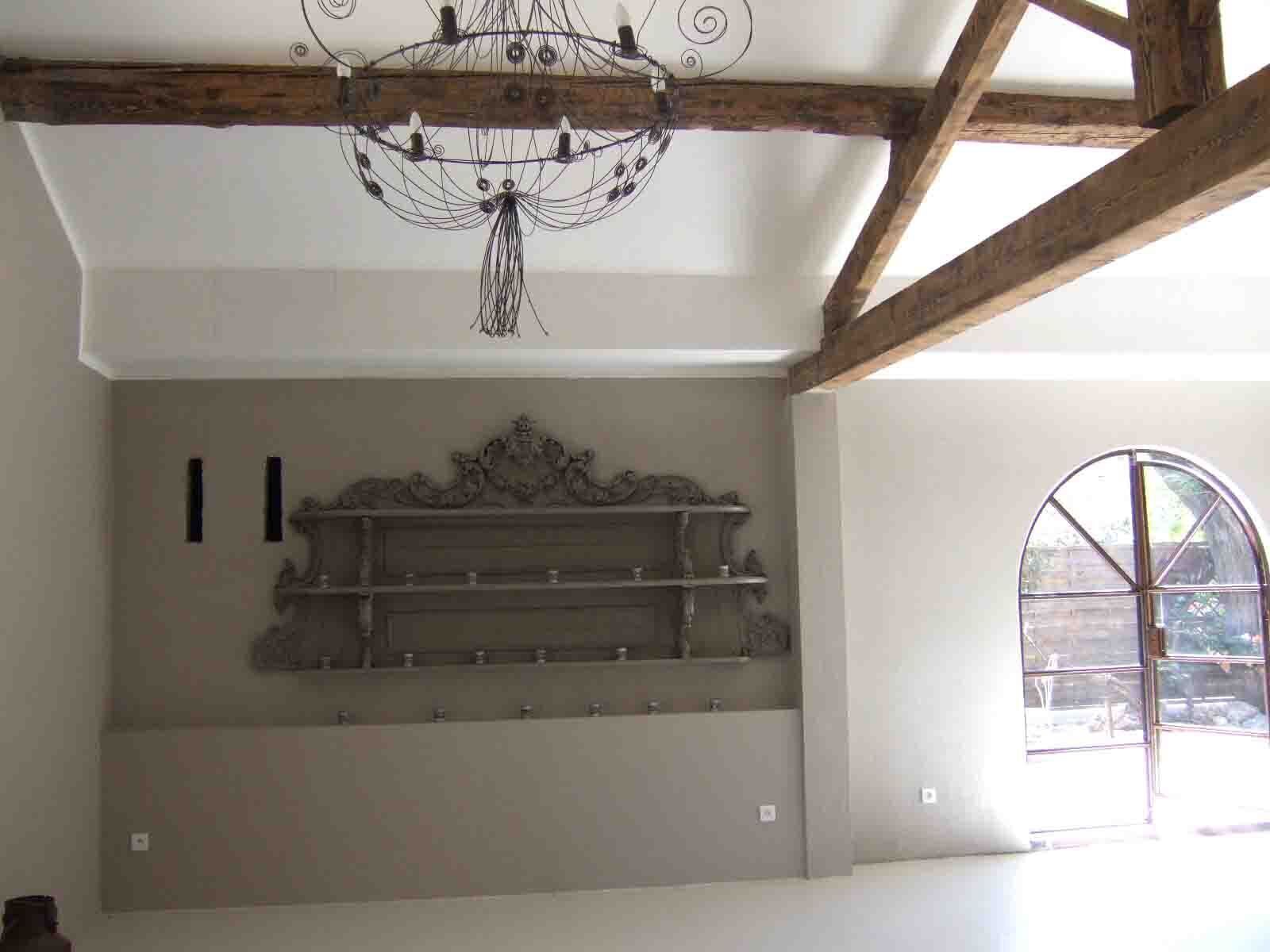 Una habitación con vigas de madera a la vista, una lámpara decorativa, una estantería fijada a la pared y un amplio ventanal arqueado.