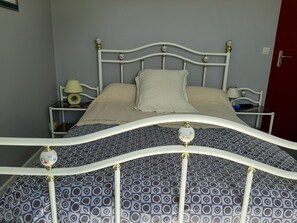 A bed with a white metal frame and a patterned bedspread. There is a nightstand with a lamp and a small table with a phone and a book.