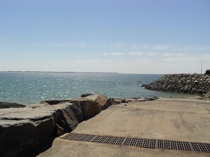 A coastal view with a concrete walkway leading to the sea, a rocky breakwater, and a distant shoreline.