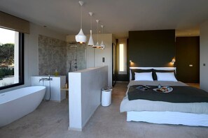 A modern hotel room with a large bathtub, a separate shower area, a bed with a dark headboard, and a view of the outdoors.