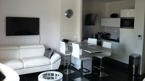 Een moderne woonkamer met een witte bank, een eettafel met stoelen, een aan de muur bevestigde televisie, en een keuken met een magnetron en spoelbak.