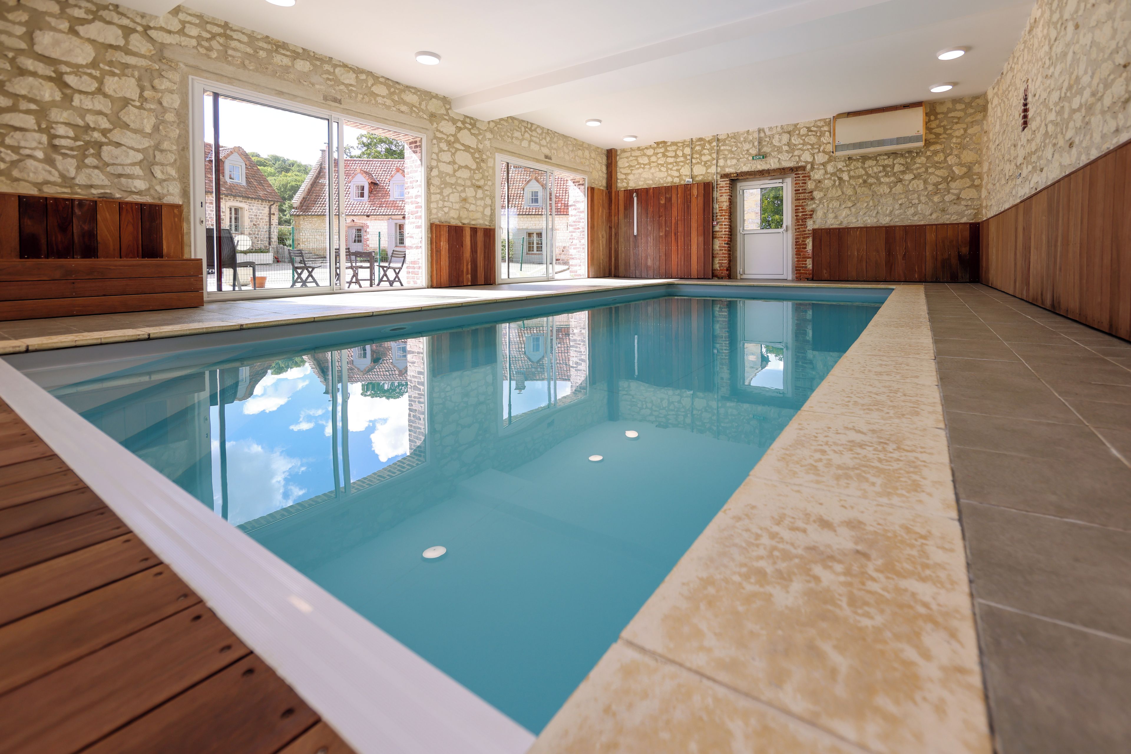 An indoor swimming pool with stone walls and wooden paneling, large windows, and a view of the outside.