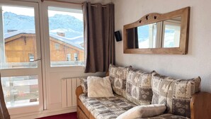 Ein gemütliches Zimmer mit Holzsofa, einem großen Fenster mit Vorhängen, einem Spiegel an der Wand und einem Heizkörper unter dem Fenster.
