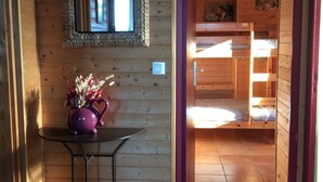 Interior de madera con piso de baldosas, una mesa pequeña con un florero, un espejo y una cama individual a la vista a través de un umbral abierto.