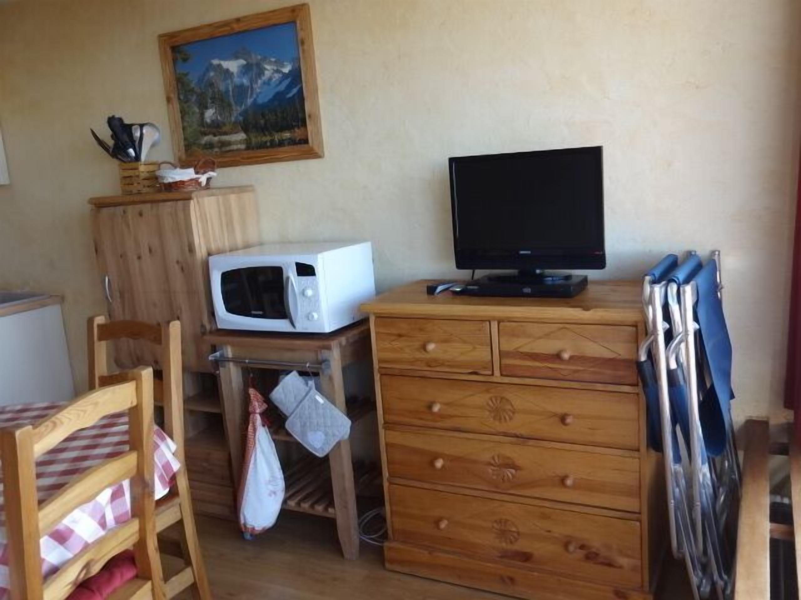 A small kitchen with a microwave, a television, a wooden cabinet, and a chair with a checkered tablecloth.