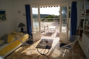 A room with a sofa, a table with a basket, two chairs, a lamp, and a view through a glass door to an outdoor patio with chairs and a railing. - NEO-PROVENCAL VILLA WITH SECURE INFINITY POOL (Roquebrune-Sur-Argens)