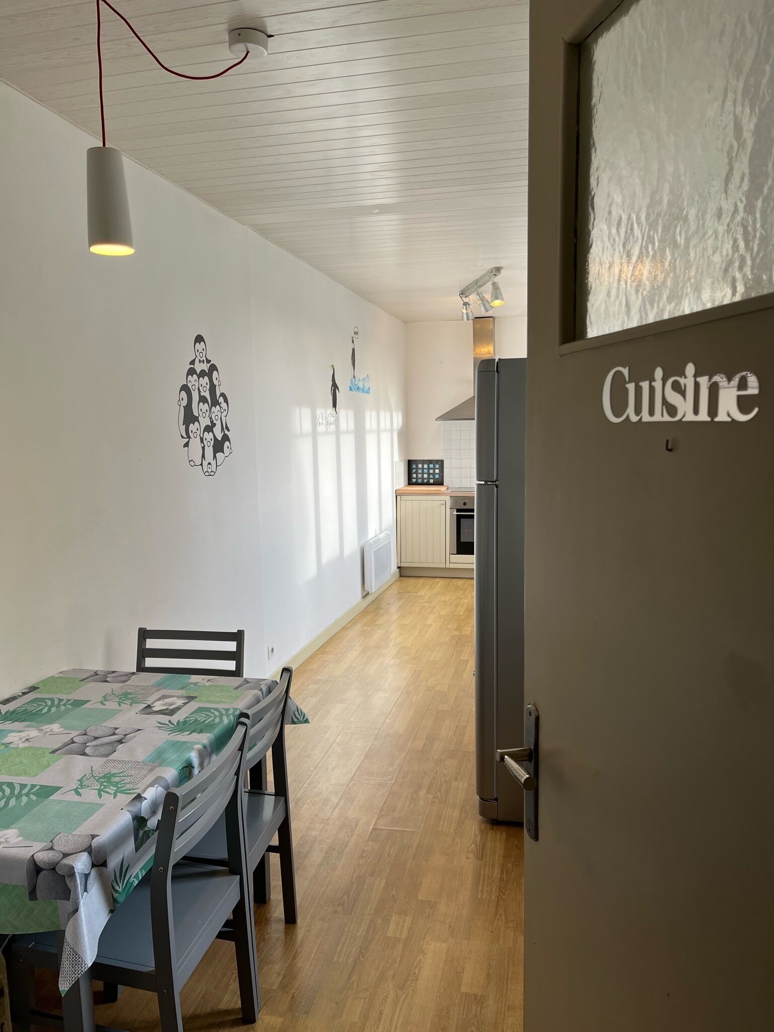 A modern kitchen with a refrigerator, a dining area with a table and chairs, and a wall with a 'Cuisine' sign.