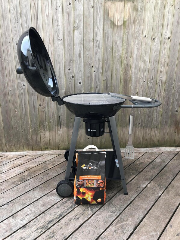 A portable grill with a lid open, a bag of charcoal, and a metal spatula on a wooden deck.