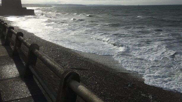 Eine Küstenlandschaft mit einem hölzernen Geländer am Strand, Wellen, die auf das Ufer branden, und einem bewölkten Himmel.