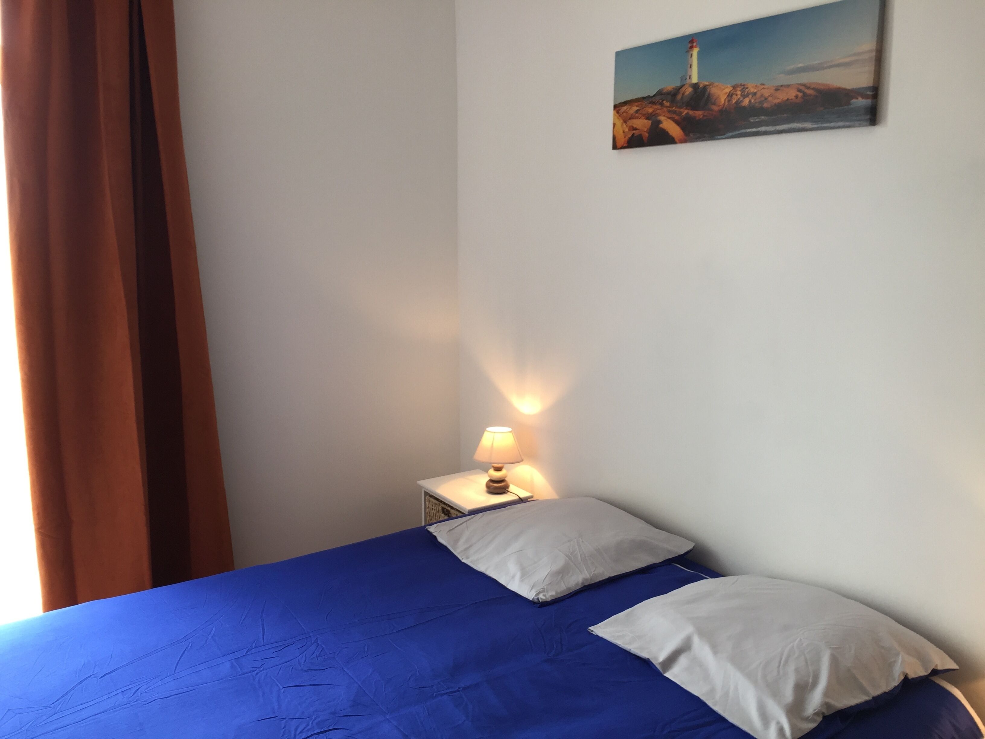 Une chambre avec un lit, une table de chevet avec une lampe, un tableau au mur et une fenêtre avec des rideaux.