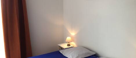 Une chambre avec un lit, une table de chevet avec une lampe, un tableau au mur et une fenêtre avec des rideaux.