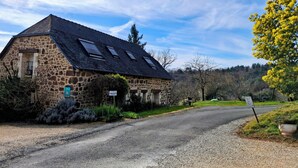 Un edificio de piedra con techo inclinado y tragaluces, rodeado de vegetación y un sendero de grava.