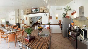 Una cocina amplia con zona de comedor, que incluye una isla central con pileta, estufa y microondas. Se observa una escalera que sube a un nivel superior, y la cocina tiene una pared de piedra rústica y un salpicón de azulejos.