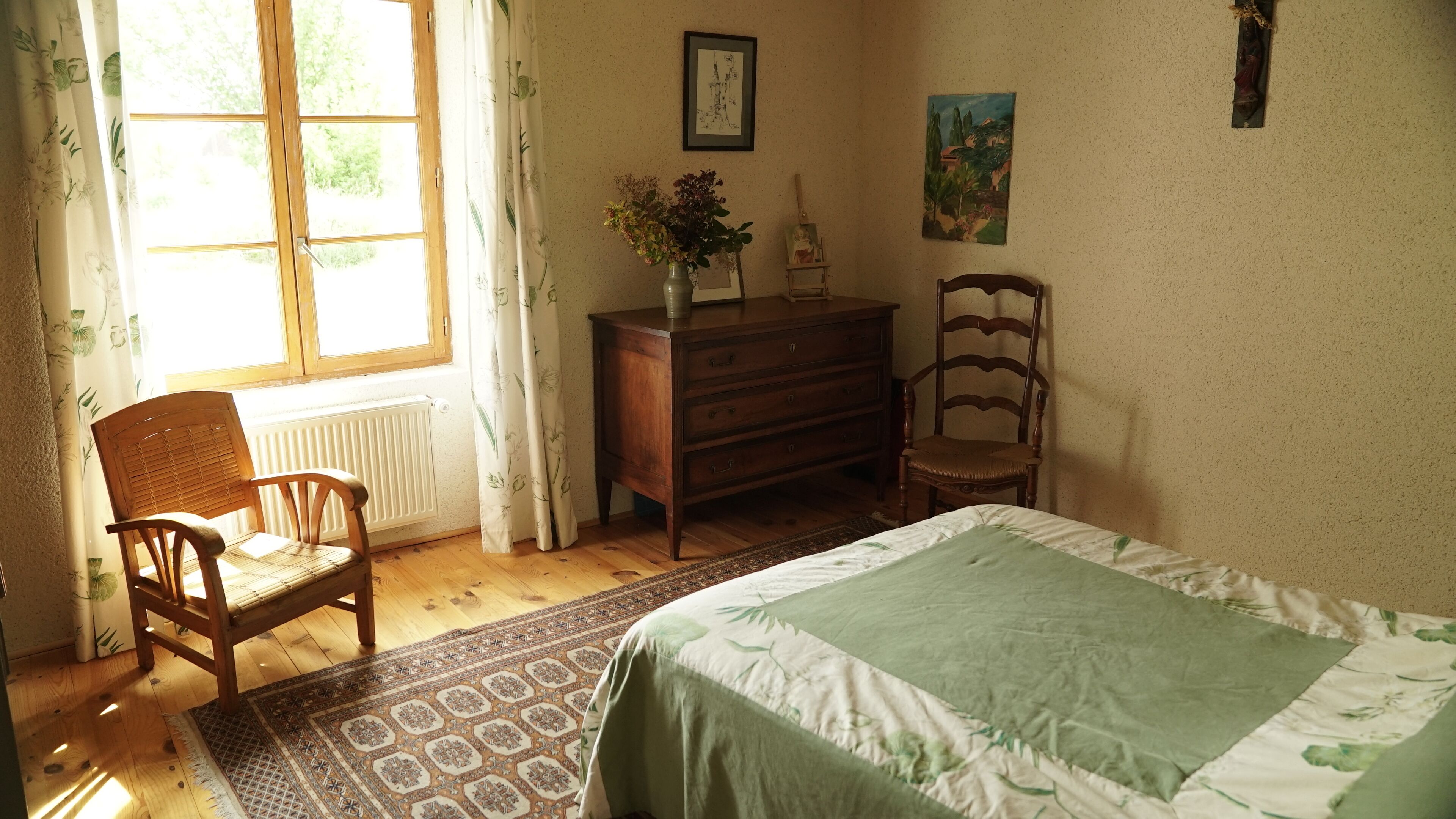 A bedroom with a bed, two wooden chairs, a dresser, a radiator, and a window with curtains.