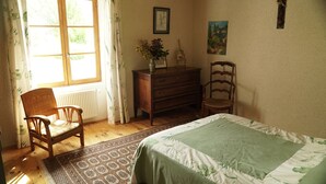 Une chambre à coucher avec un lit, deux chaises en bois, une commode, un radiateur et une fenêtre avec des rideaux.