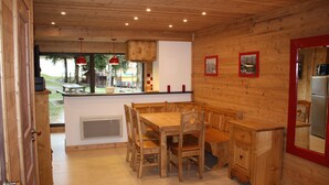 Een houten interieur met een eettafel en stoelen, een keukenhoek met een koelkast, en uitzicht door een raam.