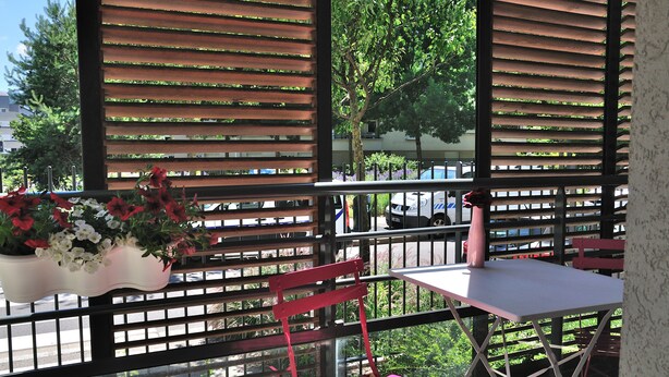 A balcony with a table and chairs, a potted plant with red and white flowers, and a view of the street and trees. - Furnished studio, near beach and town center, WIFI  (Annecy)