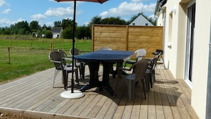 Une terrasse en bois avec une table et des chaises, un parasol et une vue sur une cour clôturée et des bâtiments en arrière-plan.