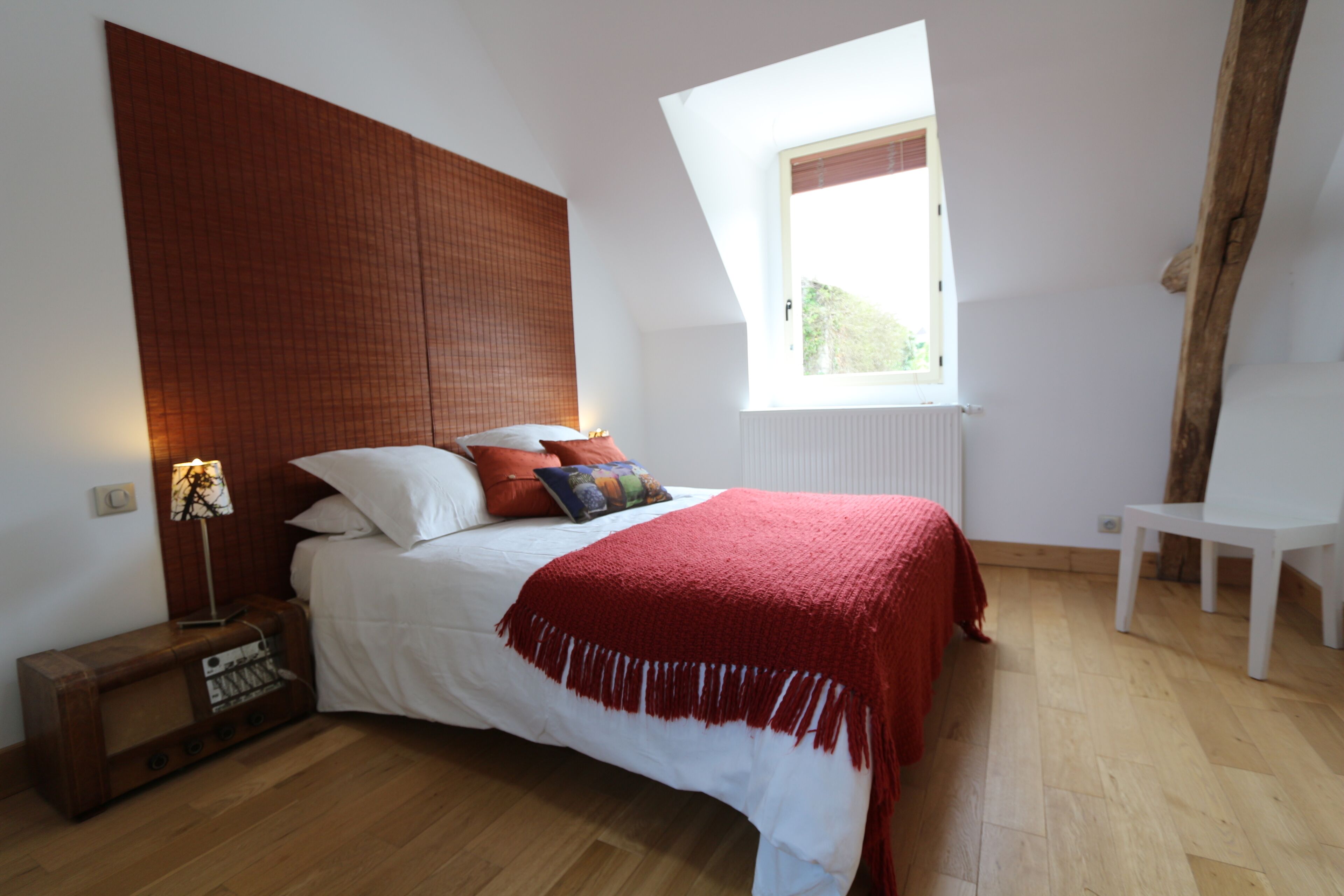 Een slaapkamer met een bed, nachtkastje, lamp, raam en stoel.