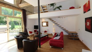 Een ruime woonkamer met een rode bank, zwarte fauteuils en een houten trap die naar een verdieping leidt.