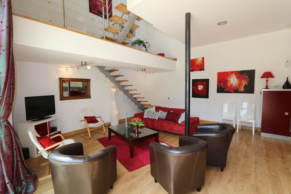 A modern living room with a staircase, red sofa, leather armchairs, and a flat-screen TV.