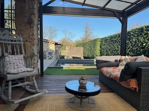Eine überdachte Terrasse mit Holzboden, einem Korbssessel, einem Schaukelstuhl und einem kleinen Tisch mit einer Pflanzen-Schale.