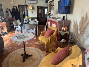 Ein gemütliches Wohnzimmer mit Fliesenboden, einem runden Tisch mit einem Buch, zwei gelben Sessel mit roten Kissen, einer Holzkommode und einem an der Wand montierten Fernseher.