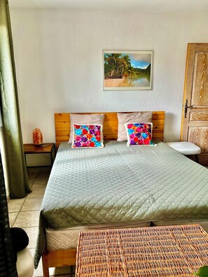 Um quarto com uma cama, duas almofadas coloridas, um quadro de uma praia, uma porta de madeira e uma mesa de cabeceira com uma luminária.