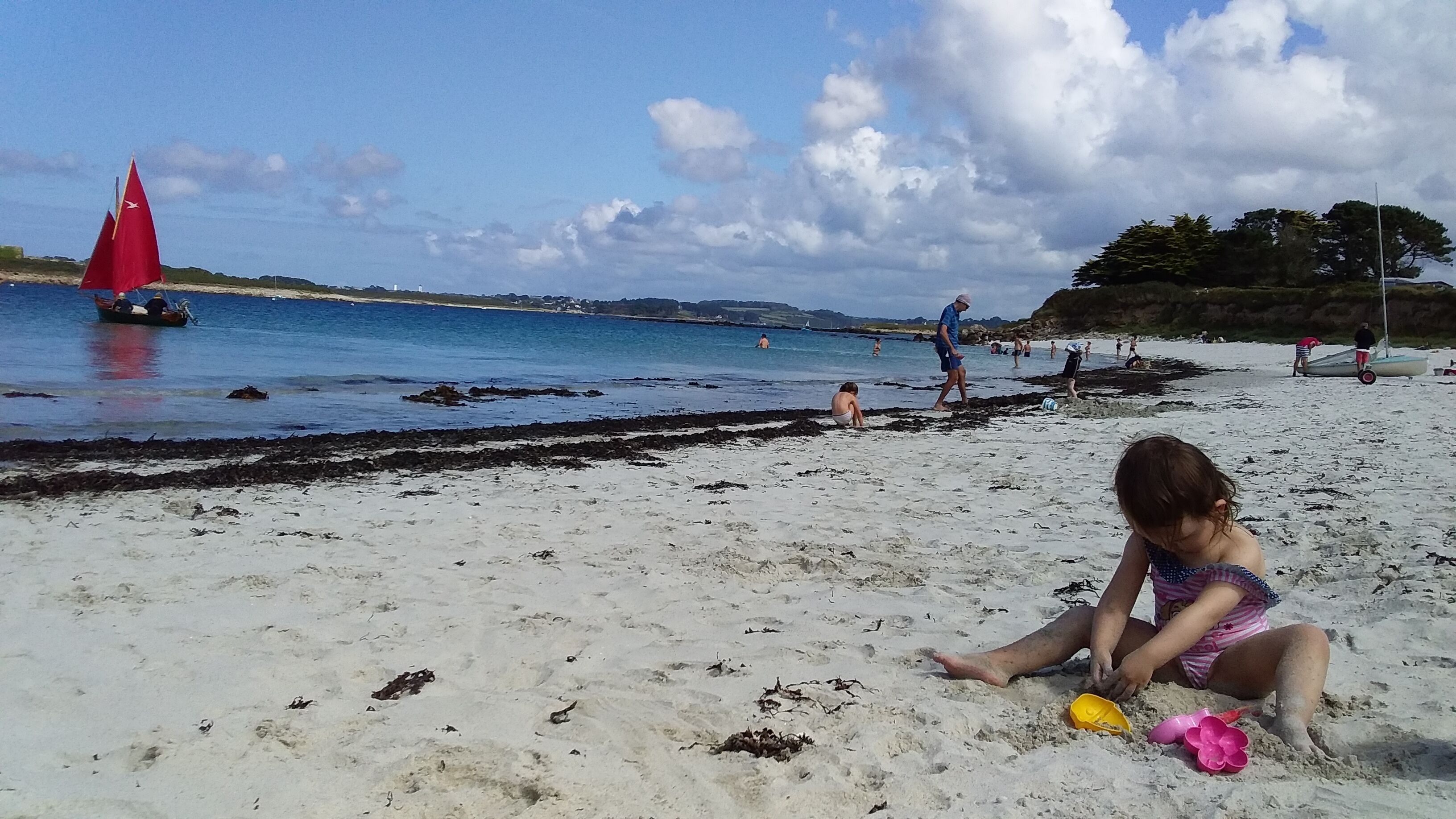 Une plage de sable avec un voilier sur l’eau, des gens qui nagent et qui se promènent, et un enfant qui joue dans le sable.
