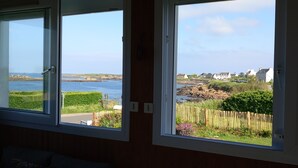 Ein Zimmer mit drei Fenstern und Blick auf eine Küstenlandschaft, inklusive Meer, felsigem Ufer und Wohnhäusern.