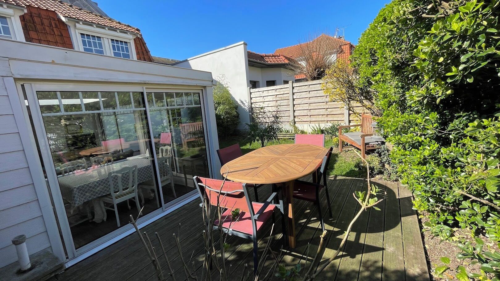 Une terrasse couverte avec une table ronde en bois et des chaises rouges, entourée d’une clôture en bois et de verdure.